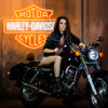 Harley Davidson neon sign in the colors orange and white. Installed on wall. Infront of neon sign is a beautiful girl posing with her black motorbike. 
